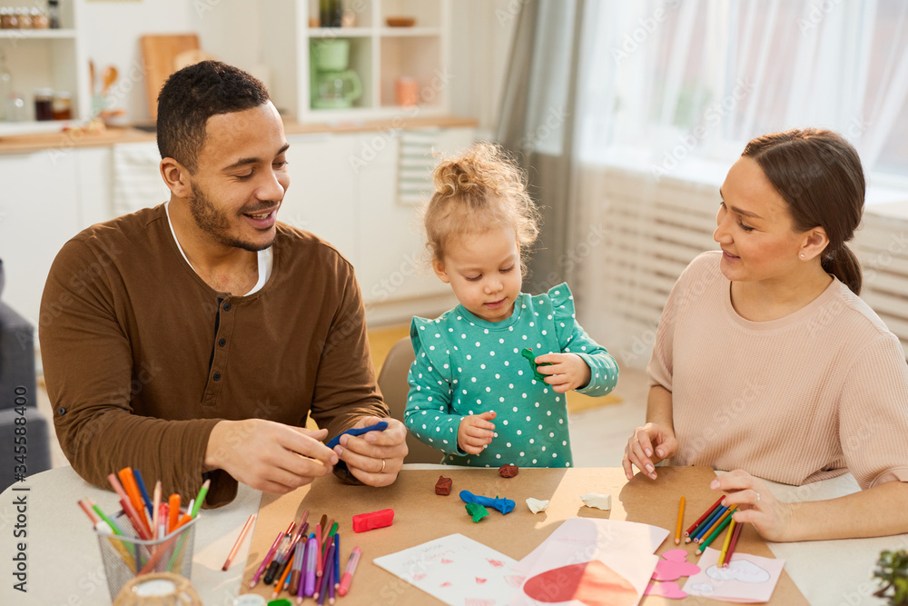 Fototapeta premium Young adult parents spending with their cute little daughter sitting together at table making models using colourful play dough