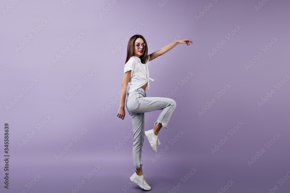 Slim girl in cute white sneakers posing on one leg in studio. Indoor photo of dreamy woman in jeans dancing on bright purple background.