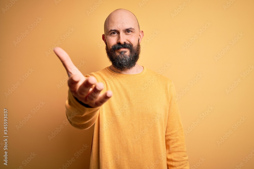 Handsome bald man with beard wearing casual sweater standing over yellow background smiling friendly offering handshake as greeting and welcoming. Successful business. Handsome bald man with beard wearing casual sweater standing over yellow background smiling friendly offering handshake as greeting and welcoming. Successful business.