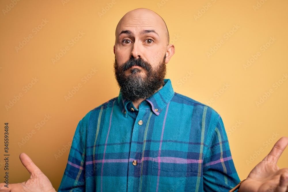 Fototapeta premium Handsome bald man with beard wearing casual shirt standing over isolated yellow background clueless and confused expression with arms and hands raised. Doubt concept.