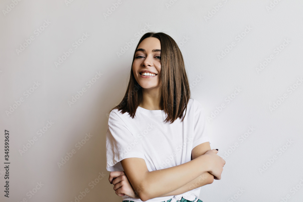 Obraz premium Pleasant girl with shiny brown hair smiling on white background. Indoor photo of caucasian lady standing in studio with arms folded.
