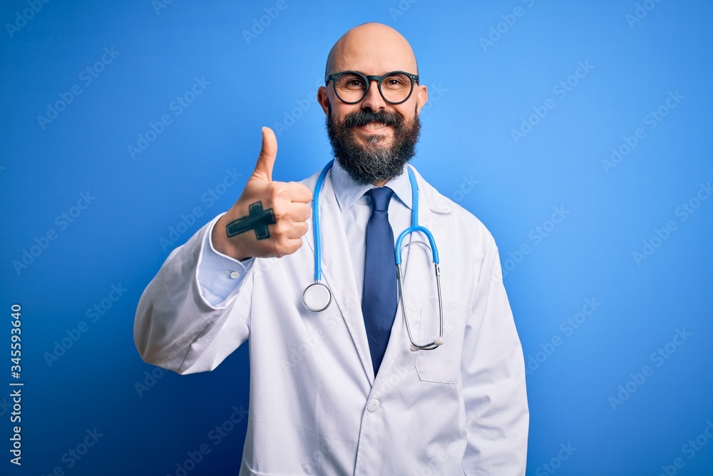 Handsome bald doctor man with beard wearing glasses and stethoscope over blue background doing happy thumbs up gesture with hand. Approving expression looking at the camera showing success. Handsome bald doctor man with beard wearing glasses and stethoscope over blue background doing happy thumbs up gesture with hand. Approving expression looking at the camera showing success.