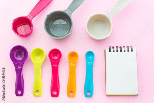 Set of sweet pastel measuring spoons, measuring cups and notepad on pink background in top view shot