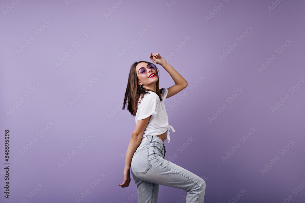 Fascinating young woman with straight hairstyle dancing in studio. Glamorous female model wears jeans and t-shirt expressing happiness on purple background. Fascinating young woman with straight hairstyle dancing in studio. Glamorous female model wears jeans and t-shirt expressing happiness on purple background.