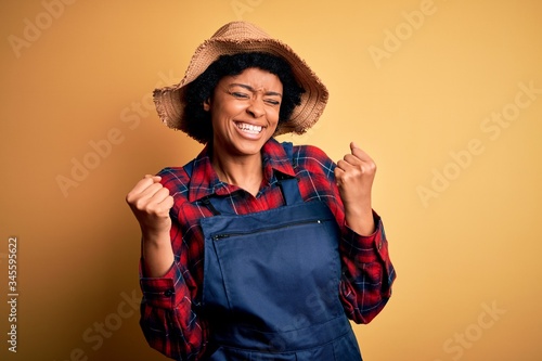 Young African American afro farmer woman with curly hair wearing apron and hat very happy and excited doing winner gesture with arms raised, smiling and screaming for success. Celebration concept.