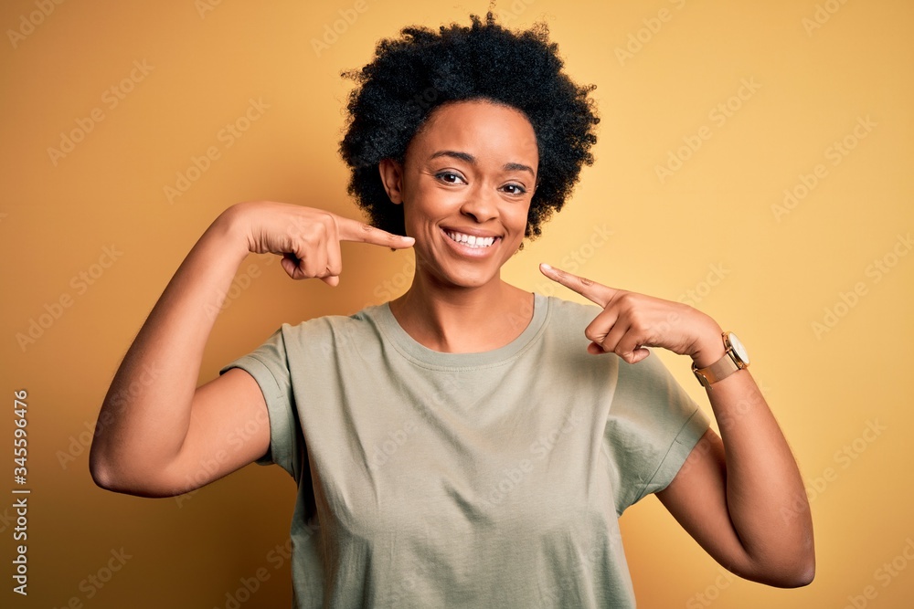 Young beautiful African American afro woman with curly hair wearing casual t-shirt smiling cheerful showing and pointing with fingers teeth and mouth. Dental health concept. Young beautiful African American afro woman with curly hair wearing casual t-shirt smiling cheerful showing and pointing with fingers teeth and mouth. Dental health concept.
