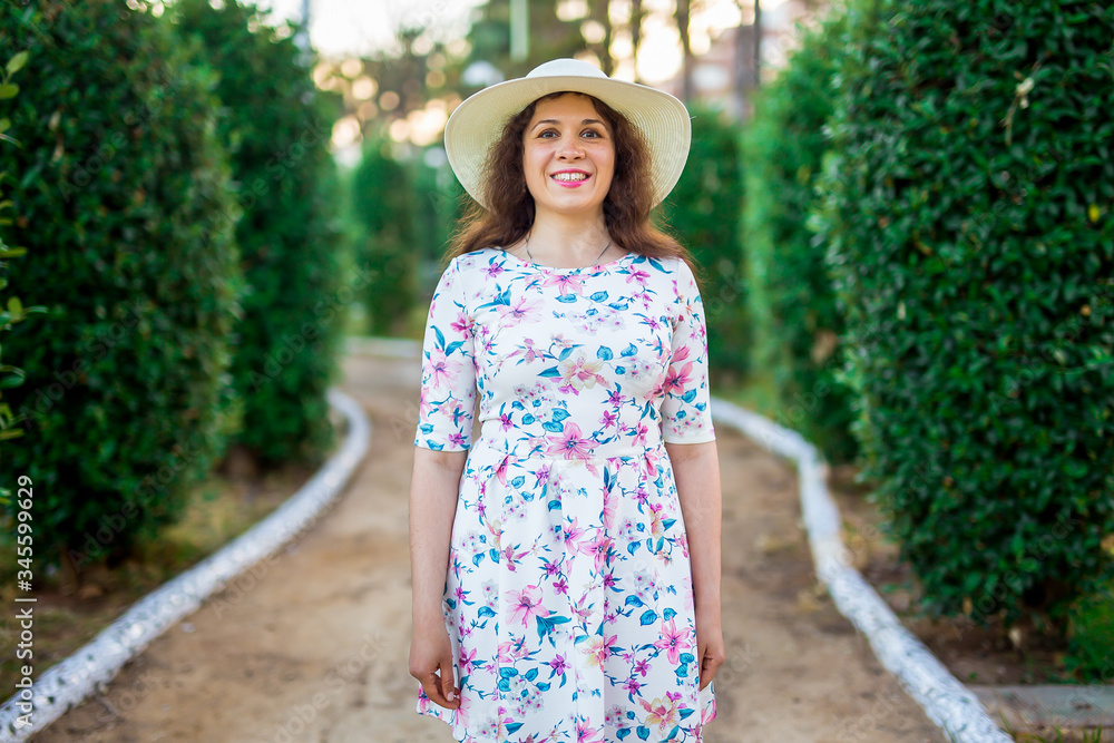 Portrait of pretty woman wearing white dress and straw hat in sunny warm weather day. Walking at summer park.