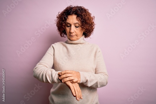 Middle age beautiful curly hair woman wearing casual turtleneck sweater over pink background Checking the time on wrist watch, relaxed and confident © Krakenimages.com