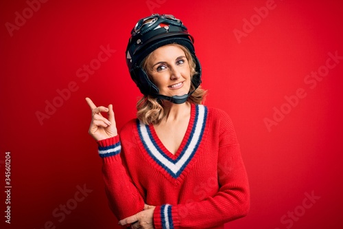 Middle age beautiful blonde motorcyclist woman wearing moto helmet over red background with a big smile on face, pointing with hand and finger to the side looking at the camera.