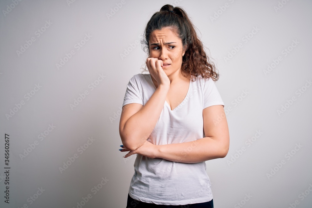Young beautiful woman with curly hair wearing casual t-shirt standing over white background looking stressed and nervous with hands on mouth biting nails. Anxiety problem. Young beautiful woman with curly hair wearing casual t-shirt standing over white background looking stressed and nervous with hands on mouth biting nails. Anxiety problem.