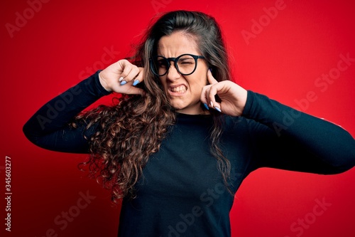 Young beautiful woman with curly hair wearing sweater and glasses over red background covering ears with fingers with annoyed expression for the noise of loud music. Deaf concept.