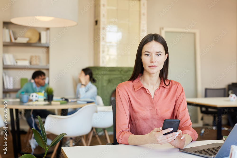 Good-looking young Caucasian woman wearing pink blouse sitting at office table holding smartphone looking at camera Good-looking young Caucasian woman wearing pink blouse sitting at office table holding smartphone looking at camera