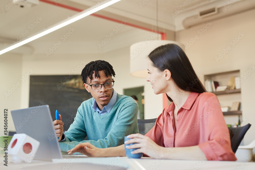 Horizontal shot of young African American man and Caucasian woman sitting together at office desk working together Horizontal shot of young African American man and Caucasian woman sitting together at office desk working together