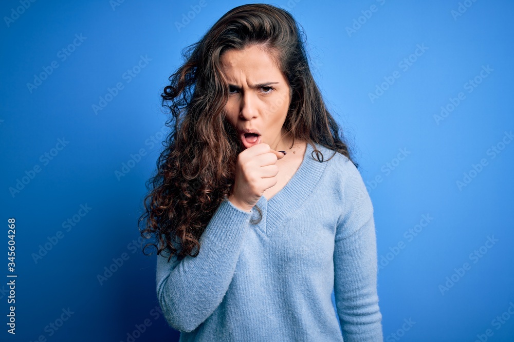 Young beautiful woman with curly hair wearing blue casual sweater over isolated background feeling unwell and coughing as symptom for cold or bronchitis. Health care concept. Young beautiful woman with curly hair wearing blue casual sweater over isolated background feeling unwell and coughing as symptom for cold or bronchitis. Health care concept.