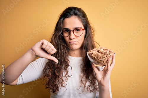 Young beautiful woman with curly hair holding bowl with healthy corn flakes cereals with angry face  negative sign showing dislike with thumbs down  rejection concept