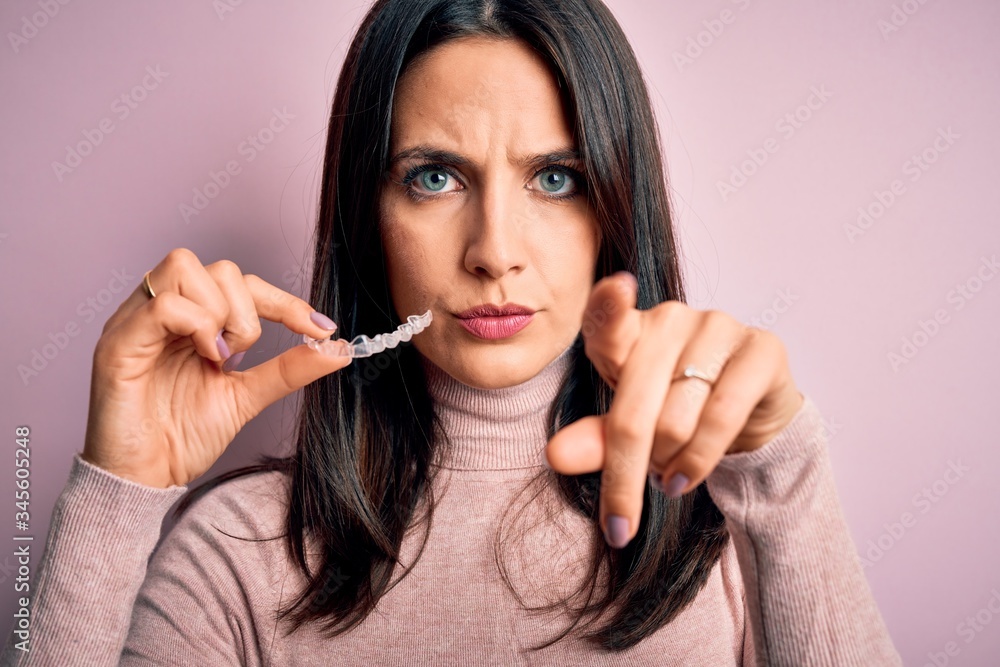 Young woman with blue eyes holding clear aligner standing over pink background pointing with finger to the camera and to you, hand sign, positive and confident gesture from the front Young woman with blue eyes holding clear aligner standing over pink background pointing with finger to the camera and to you, hand sign, positive and confident gesture from the front