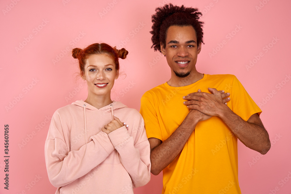 Obraz premium Kind hearted friendly looking young red haired woman and black man keep both palms on chest, dressed casually, express gratitude and good feeling, stand next to each other, over pink studio background