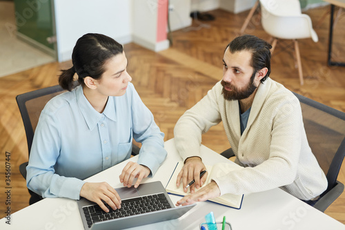 Young Caucasian man with beard on face and attractive young woman sitting together at office desk working on project, high angle view shot
