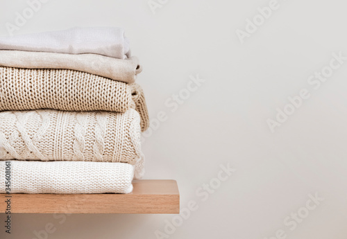 A stack of neatly folded warm knitwear, wool on a wooden shelf and a beige background. Capsule wardrobe, clothes storage, minimalism, knitted texture, order, comfort. Banner with copy space for text.