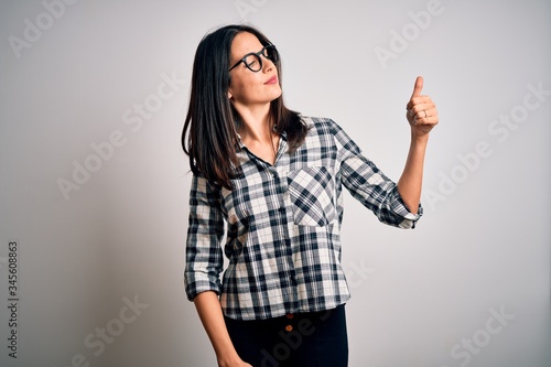 Young brunette woman with blue eyes wearing casual shirt and glasses over white background Looking proud, smiling doing thumbs up gesture to the side
