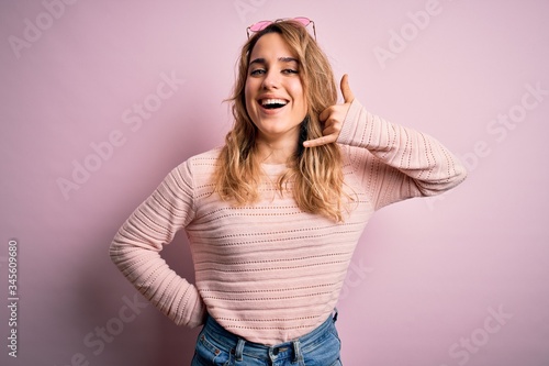 Young beautiful blonde woman wearing casual sweater and sunglasses over pink background smiling doing phone gesture with hand and fingers like talking on the telephone. Communicating concepts.