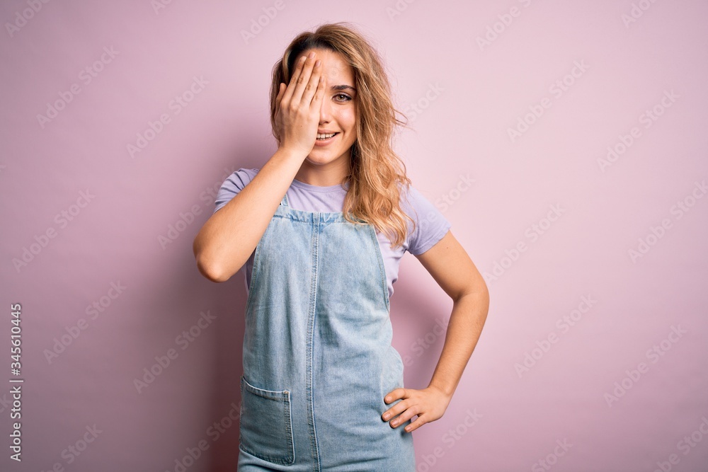 Young beautiful blonde woman wearing casual denim overalls standing over pink background covering one eye with hand, confident smile on face and surprise emotion. Young beautiful blonde woman wearing casual denim overalls standing over pink background covering one eye with hand, confident smile on face and surprise emotion.