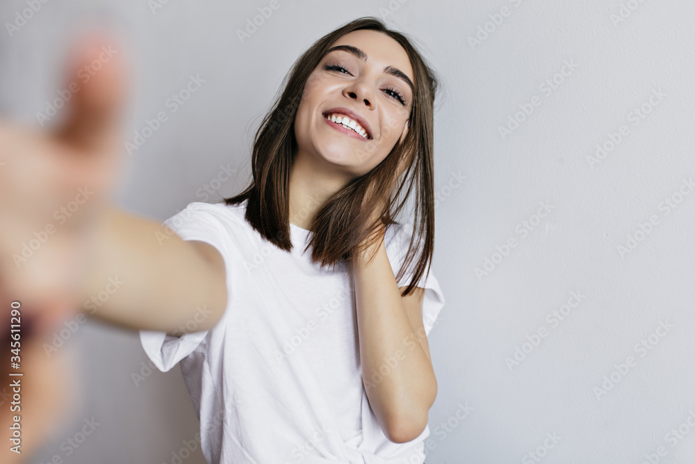 Brunette excited girl in white t-shirt making selfie. Indoor shot of emotional caucasian young lady isolated on light background. Brunette excited girl in white t-shirt making selfie. Indoor shot of emotional caucasian young lady isolated on light background.