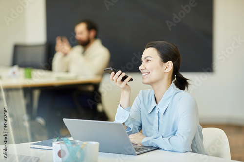 Young adult woman sitting at office table in front of laptop recording voice massage using her smartphone