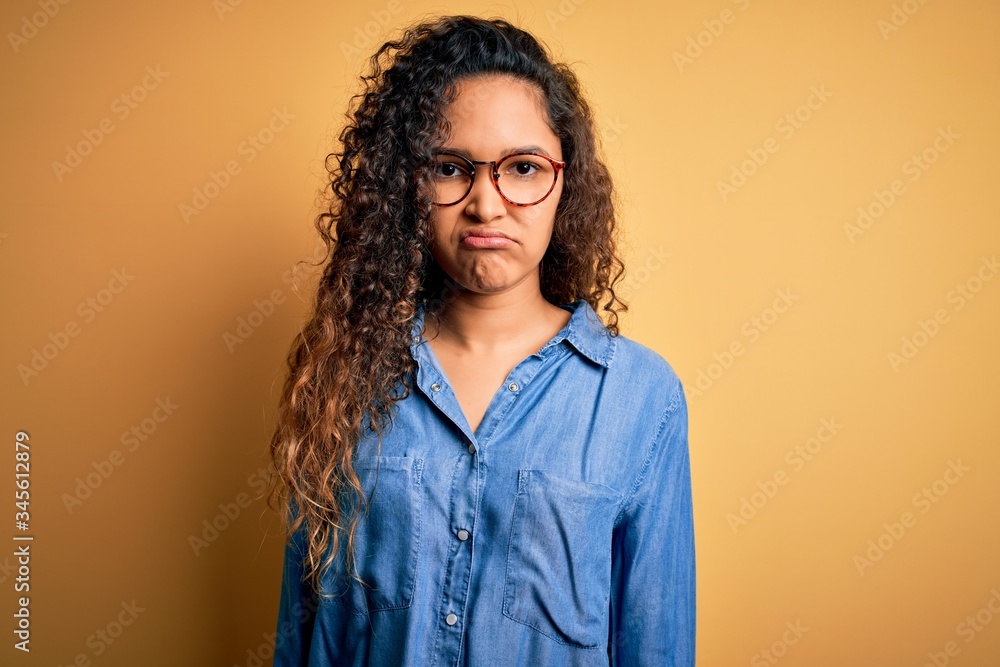Beautiful woman with curly hair wearing casual denim shirt and glasses over yellow background depressed and worry for distress, crying angry and afraid. Sad expression. Beautiful woman with curly hair wearing casual denim shirt and glasses over yellow background depressed and worry for distress, crying angry and afraid. Sad expression.