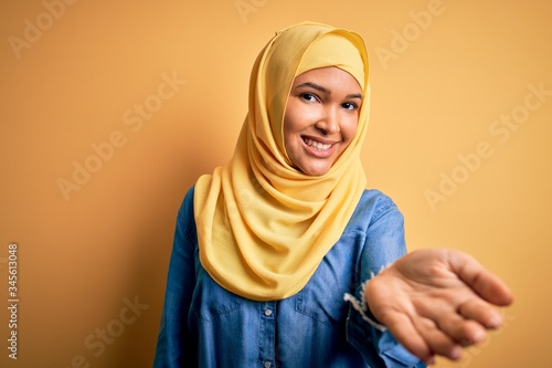 Young beautiful woman with curly hair wearing arab traditional hijab over yellow background smiling friendly offering handshake as greeting and welcoming. Successful business. © Krakenimages.com