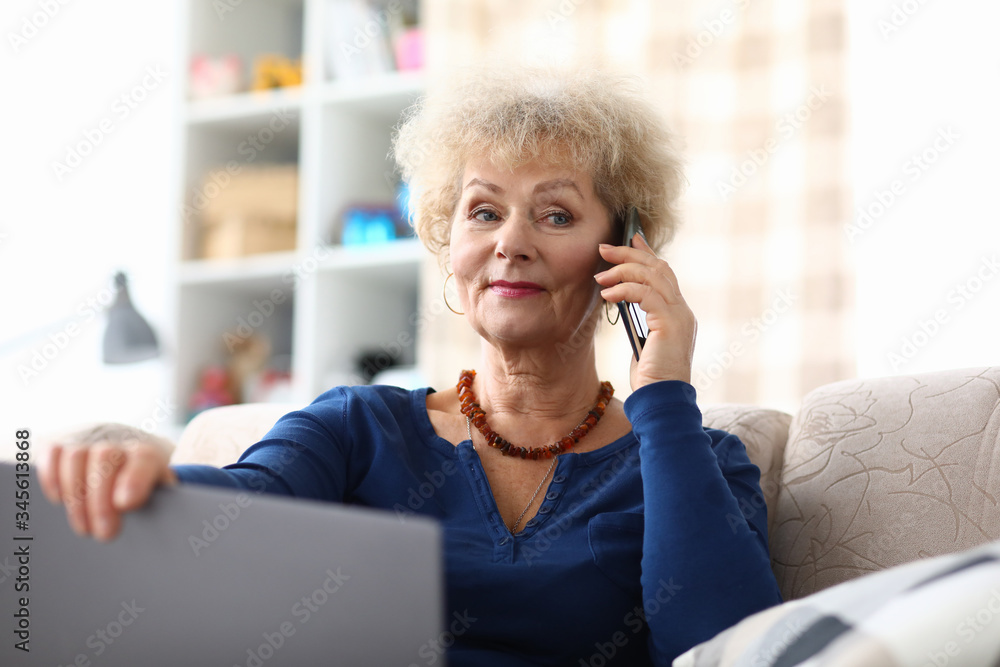 Portrait of beautiful elderly woman talking on mobile phone at home. Businesswoman holding laptop and sitting on cozy sofa. Interesting communication. Technology and leisure concept Portrait of beautiful elderly woman talking on mobile phone at home. Businesswoman holding laptop and sitting on cozy sofa. Interesting communication. Technology and leisure concept