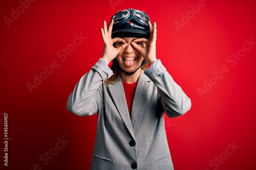 Young beautiful blonde motorcyclist woman wearing motorcycle helmet over red background doing ok gesture like binoculars sticking tongue out, eyes looking through fingers. Crazy expression.