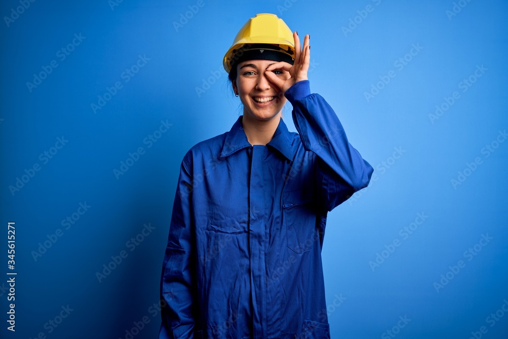 Young beautiful worker woman with blue eyes wearing security helmet and uniform doing ok gesture with hand smiling, eye looking through fingers with happy face.