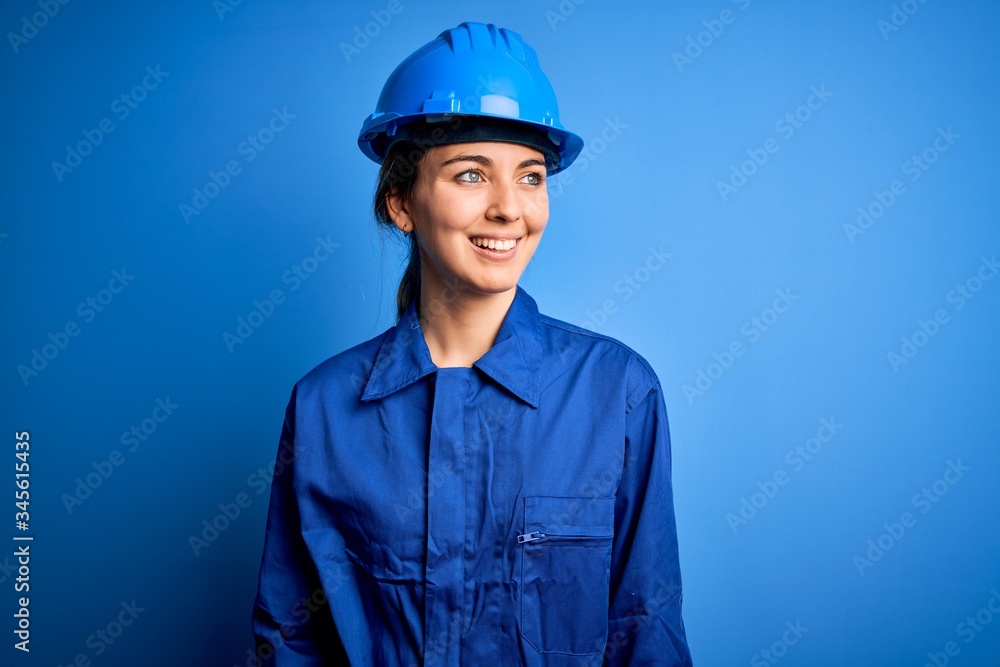 Young beautiful worker woman with blue eyes wearing security helmet and uniform looking away to side with smile on face, natural expression. Laughing confident.