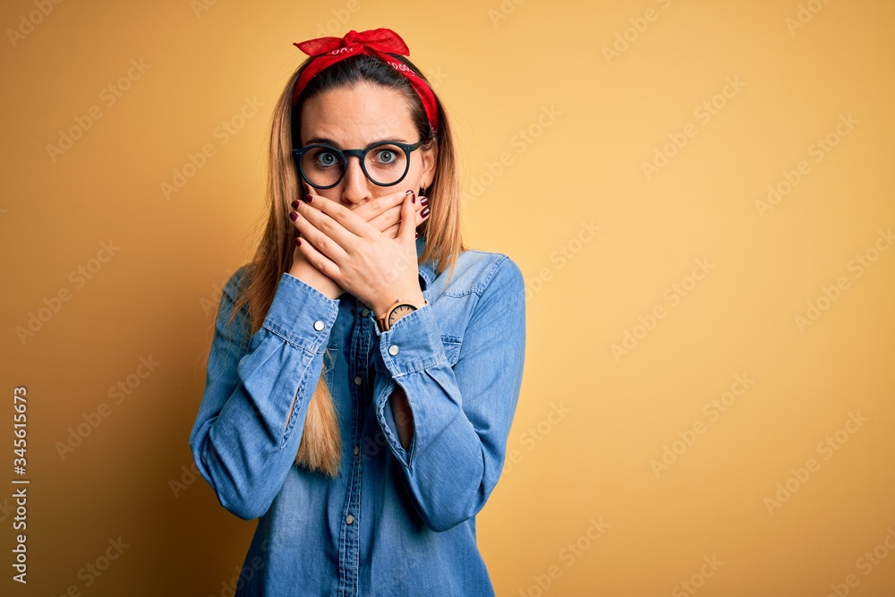 Young beautiful blonde woman with blue eyes wearing denim shirt over yellow background shocked covering mouth with hands for mistake. Secret concept. Young beautiful blonde woman with blue eyes wearing denim shirt over yellow background shocked covering mouth with hands for mistake. Secret concept.