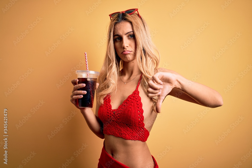 Young beautiful blonde woman on vacation wearing bikini drinking glass of fizzy cola beverage with angry face, negative sign showing dislike with thumbs down, rejection concept Young beautiful blonde woman on vacation wearing bikini drinking glass of fizzy cola beverage with angry face, negative sign showing dislike with thumbs down, rejection concept