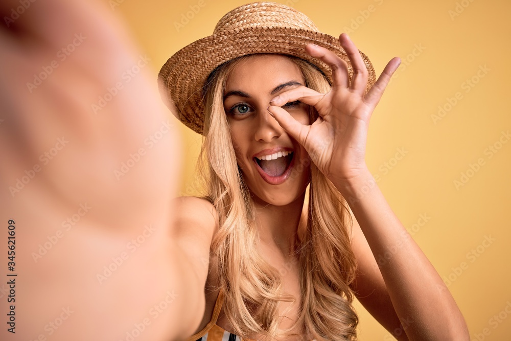 Young beautiful blonde woman on vacation wearing bikini and hat making selfie by camera with happy face smiling doing ok sign with hand on eye looking through fingers Young beautiful blonde woman on vacation wearing bikini and hat making selfie by camera with happy face smiling doing ok sign with hand on eye looking through fingers