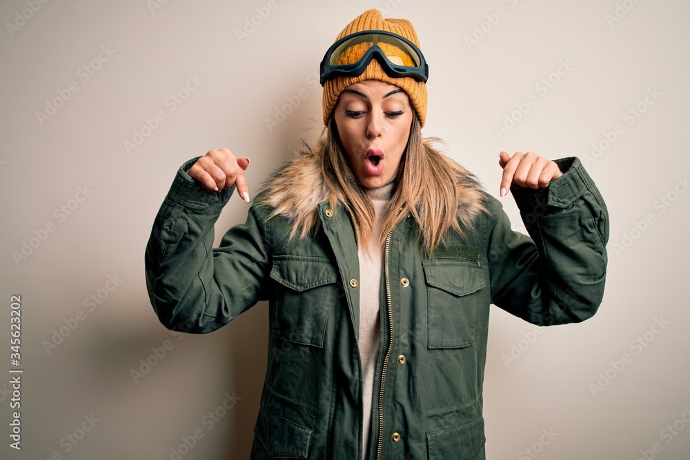 Young brunette skier woman wearing snow clothes and ski goggles over white background Pointing down with fingers showing advertisement, surprised face and open mouth Young brunette skier woman wearing snow clothes and ski goggles over white background Pointing down with fingers showing advertisement, surprised face and open mouth