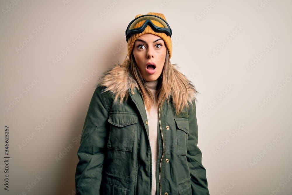 Young brunette skier woman wearing snow clothes and ski goggles over white background In shock face, looking skeptical and sarcastic, surprised with open mouth