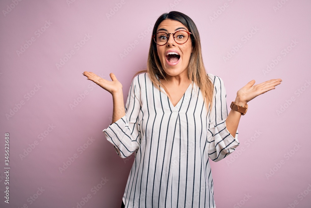 Young beautiful woman wearing casual striped t-shirt and glasses over pink background celebrating crazy and amazed for success with arms raised and open eyes screaming excited. Winner concept Young beautiful woman wearing casual striped t-shirt and glasses over pink background celebrating crazy and amazed for success with arms raised and open eyes screaming excited. Winner concept