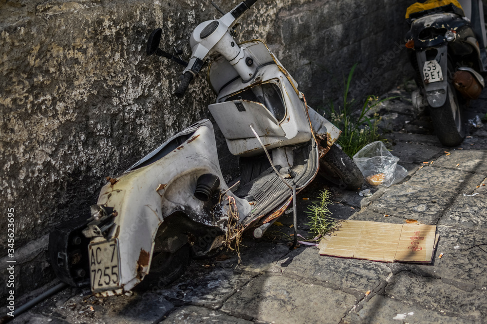 Wreck of white scooter, rusted, damaged, abandoned next to wall. Poor district of Napoli, city full of scooters and motorcycles. Urban street view, typical for slums. 
