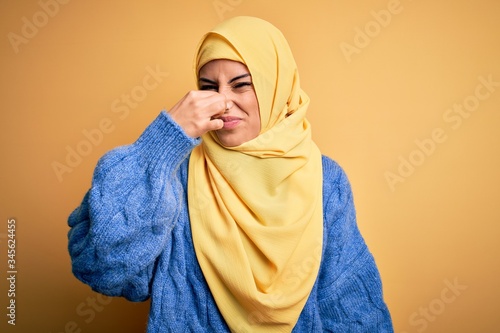 Young beautiful brunette muslim woman wearing arab hijab over isolated yellow background smelling something stinky and disgusting, intolerable smell, holding breath with fingers on nose. Bad smell © Krakenimages.com