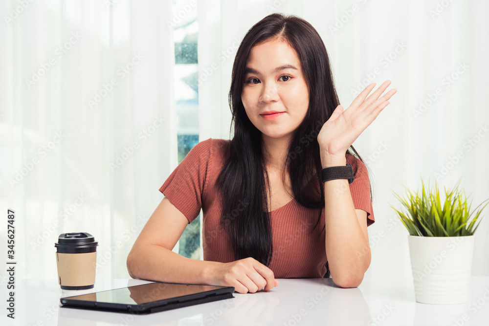 Work from home, Asian business young beautiful woman smiling sitting on desk workspace raise hand say hello communicating with team video chat or conferencing on digital tablet computer at home office Work from home, Asian business young beautiful woman smiling sitting on desk workspace raise hand say hello communicating with team video chat or conferencing on digital tablet computer at home office