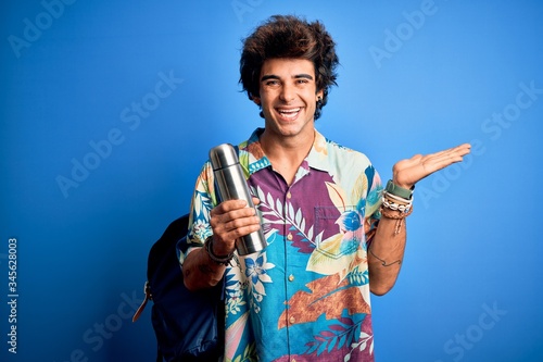 Young tourist man wearing backpack holding thermo over isolated blue background very happy and excited, winner expression celebrating victory screaming with big smile and raised hands