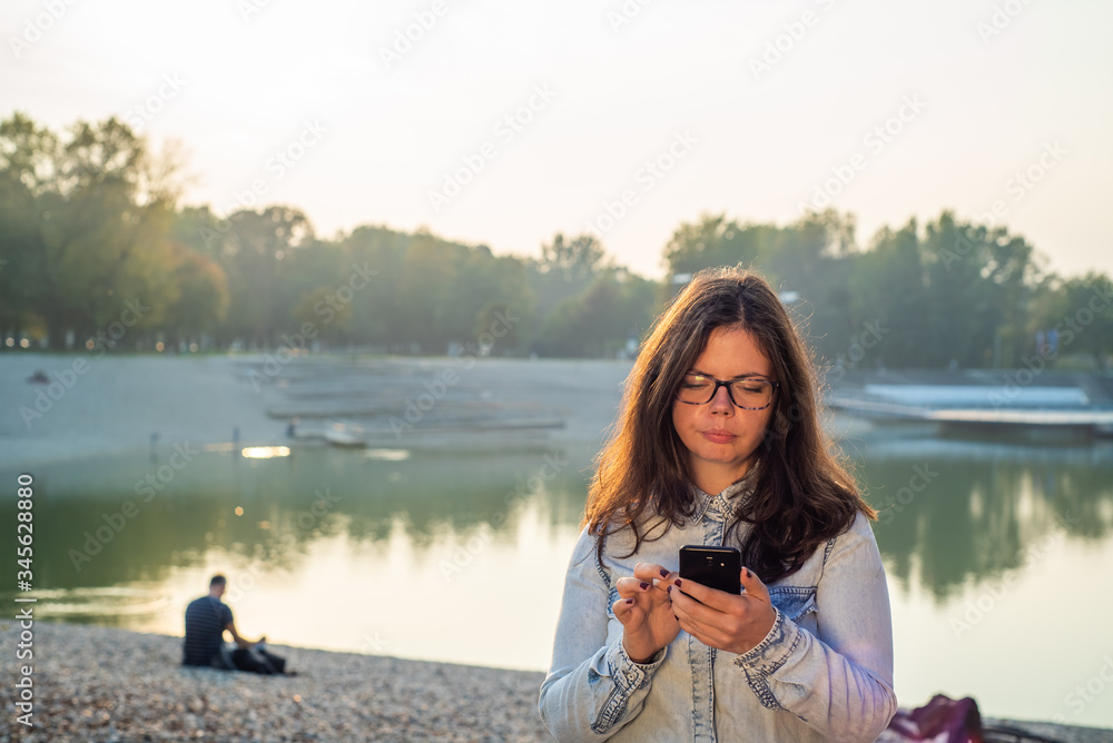 Young girl addictive to a cell phone typing in front of lake.