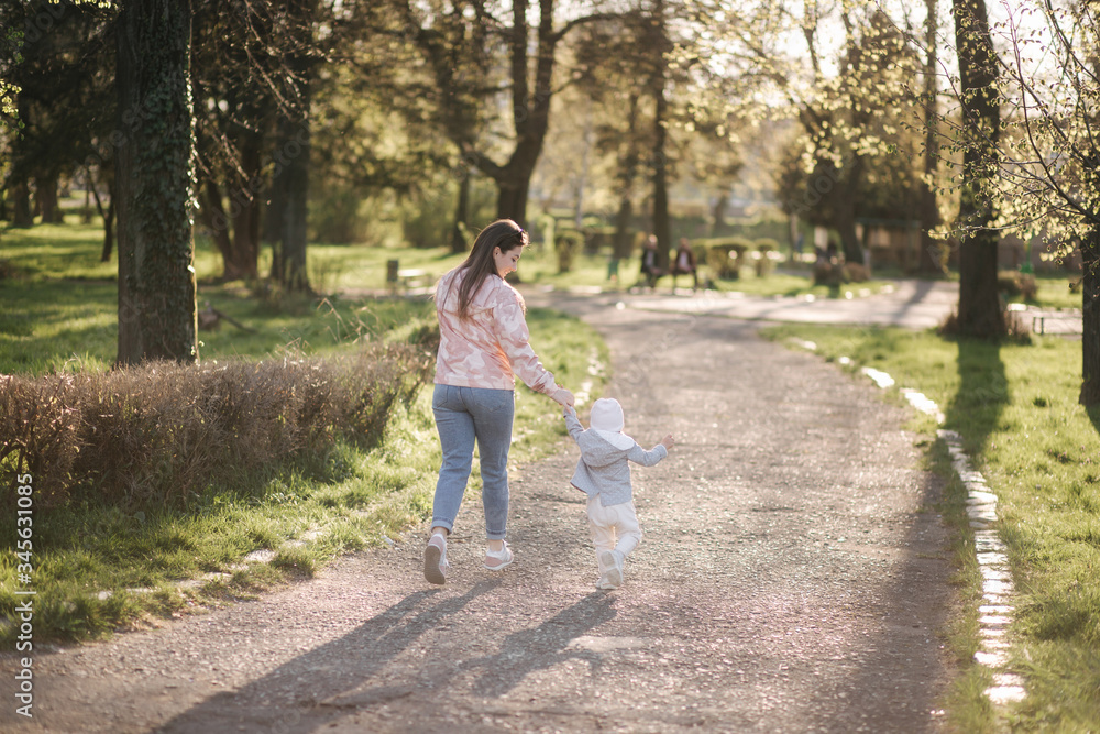 Fototapeta premium Back view of Beautiful mom with adorable daughter walk in the park. Happy family spend time in Mother's Day. Cute little girl with mother. Stylish female run and have fun