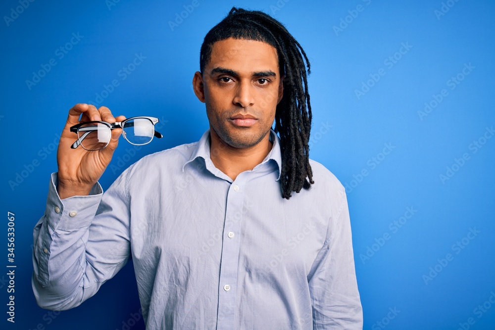 Fototapeta premium Young african american afro man with dreadlocks holding glasses over blue background with a confident expression on smart face thinking serious
