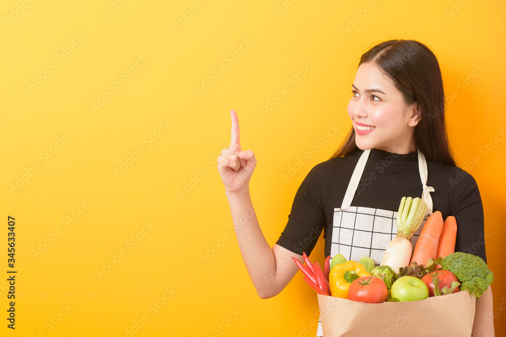 Beautiful young woman is holding vegetables in grocery bag in studio yellow background Beautiful young woman is holding vegetables in grocery bag in studio yellow background