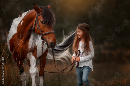 Little girl in indian style with pinto pony in a summer field in sunny evening