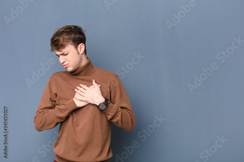 young blonde man looking sad, hurt and heartbroken, holding both hands close to heart, crying and feeling depressed isolated against flat wall photo